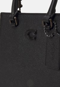 Sac à main en cuir noir texturé avec des poignées cousues, logo lettre "C" noir, et étiquette attachée à une chaîne à billes en métal.
