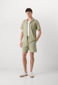 Lichtgroene short-sleeve shirt en bijpassende shorts van een gestructureerde stof met een geruit patroon. Er wordt een witte tanktop eronder gedragen.