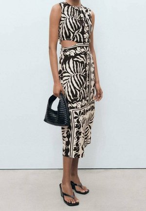 Kvinde iført en ærmeløs, sort og beige mønstret midikjole med udskæring i siden, holdende en sort flettet håndtaske og iført sorte høje sandaler.