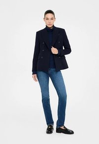 Marineblauer, doppelt geknöpfter Blazer mit Reverskragen und Seitentaschen, getragen über einem marineblauen Rollkragenpullover, kombiniert mit hellblauen Jeans und schwarzen Loafers.