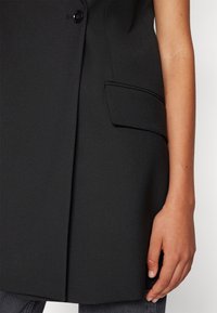 Zbliżenie na czarną, bezrękawnikową marynarkę z guzikiem i klapkową kieszenią obok ramienia i ręki osoby.