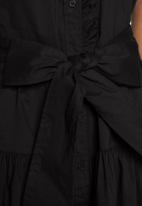 Schwarzes Kleid mit texturiertem Oberteil, gerafften Details und gebundener Taille. Verfügt über einen Knopfverschluss und einen fließenden Rock. Weiche Stofftextur.