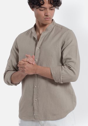 Giovane uomo con capelli ricci che indossa una camicia beige con bottoni e maniche arrotolate, guarda verso il basso, mani intrecciate davanti.