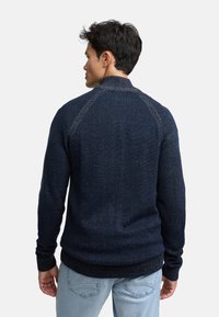 Marineblauw trui met een gestructureerd patroon, geribbelde manchetten en zoom, en een hoge kraag. Gedetailleerd stiksel langs de schouders benadrukt het ontwerp.