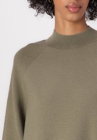 Khaki green knit sweater with a high, ribbed neckline and raglan sleeves, featuring a textured, soft fabric. Close-up view.