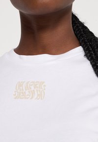 Biała bawełniana koszulka z teksturą kremowego, graficznego napisu na piersi. Projekt zawiera zdeformowane litery w jasnobeżowym kolorze.