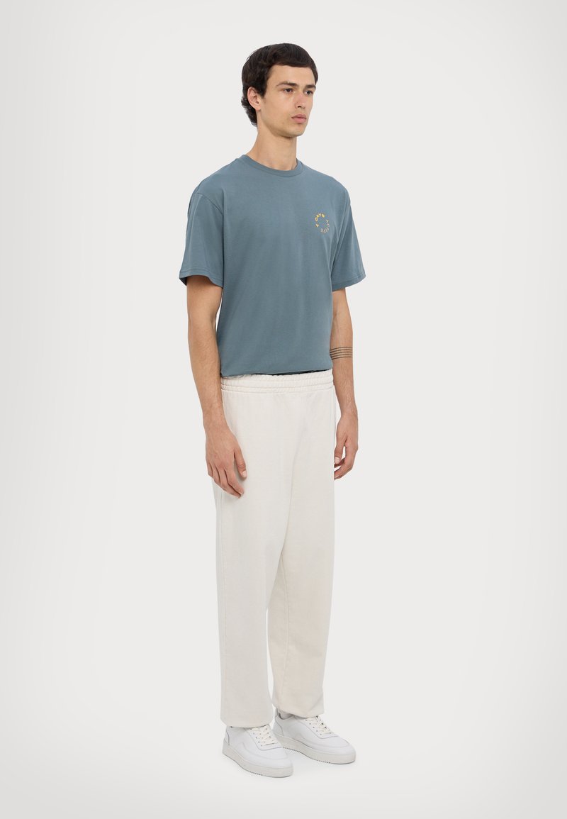 Blue cotton short-sleeve t-shirt with a round neckline and small graphic detail. Paired with loose-fitting, white pants and white sneakers.