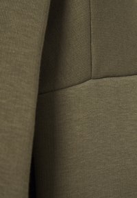 Tejido verde oliva con textura acanalada, con un diseño ajustado y costuras en contraste. Superficie lisa con un ligero brillo.