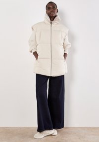Gevoerde crèmekleurige vest met hoge kraag, rits aan de voorkant, geribbelde mouwen, twee zijzakken, gestyled met wijde donkere jeans en lichte sneakers.