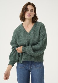 Pull en tricot vert surdimensionné avec un col en V, des poignets et un ourlet côtelés. Texture douce avec un léger éclat. Associé à un jean bleu ajusté.