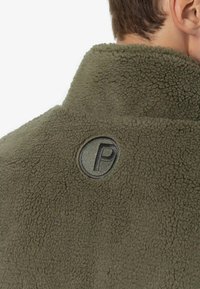 Maslinasto zelena flis jakna s teksturom mekanog, gustog materijala; ima okrugli oznaku sa utisnutim slovom "P" na leđima.