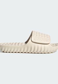 Vaalean beige liukuva sandaali, jossa on teksturoitu hihna ja pehmustettu pohja, jossa on koholla olevia pisteitä ja aaltoileva pohjan muotoilu.