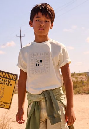 Ragazzo che indossa una maglietta bianca con la scritta "On the Road" e una giacca verde legata alla vita, in piedi vicino a un cartello di avvertimento alieno sulla strada nel deserto.