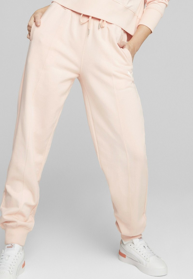 Pantalones de chándal rosa claro con cinturilla elástica y cordón. Tiene bolsillos laterales, piernas estrechas y puños de canalé. Combinados con zapatillas blancas.