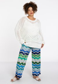 Witte gebreide trui met een chevronpatroon, gecombineerd met wijd uitlopende broek met blauwe, groene en crèmekleurige zigzagstrepen. Sandalen zijn beige.