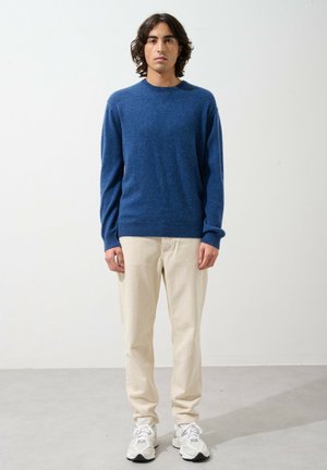 Blauer Wollpullover mit Rundhalsausschnitt und gerippten Bündchen, kombiniert mit beigen Hosen und weißen Sneakers. Minimalistisches Design mit weicher Textur.