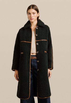 Femme portant un long manteau en laine noir à boutons avec des détails marron, un pull court beige, un jean bleu foncé et une ceinture marron sur fond beige.