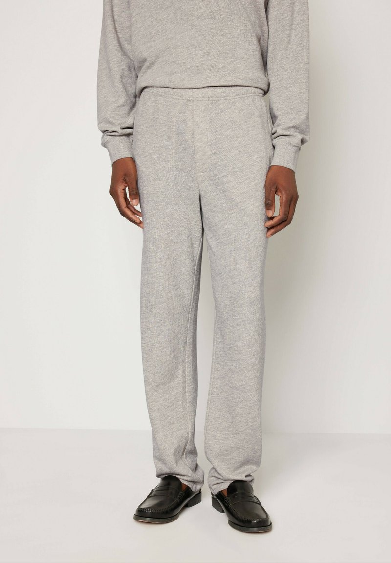 Sive pletene trenirke z elastičnim pasom in ravnim krojem, v kombinaciji s črnimi loaferji, na beli podlagi.