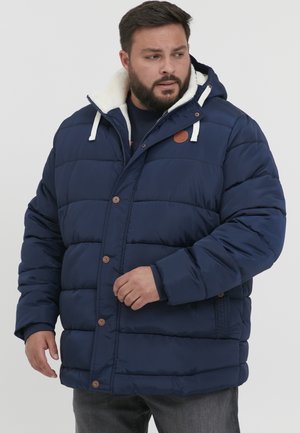 Chaqueta tipo poncho de color azul marino con capucha forrada de felpa blanca, botones a presión y bolsillos laterales. Presenta un diseño acolchado y un parche de logo en color beige.