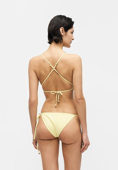Γυναίκα με κοντά σκοτεινά μαλλιά που φοράει ανοιχτό κίτρινο μπικίνι με ιμάντες που σταυρώνονται στην πλάτη και σλιπ που δένει στο πλάι, στέκεται μπροστά σε ένα απλό ανοιχτό φόντο.