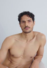 Un mannequin masculin torse nu avec des cheveux courts, foncés et ondulés, assis contre un fond gris clair, affichant des teintes de peau naturelles et une texture lisse.