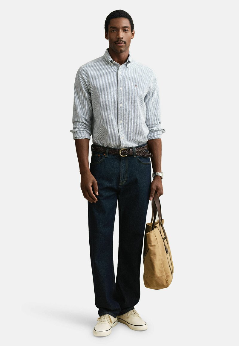 Hombre con camisa de rayas abotonada, vaqueros oscuros, zapatillas blancas, sosteniendo una bolsa de lona color beige con asas marrones, de pie contra un fondo blanco.