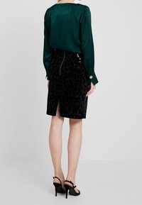 Blusa de seda verde con una textura suave y hombros plisados, combinada con una falda de lápiz negra adornada con lentejuelas y con una abertura en la parte trasera. Tacones altos negros.