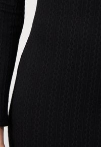 Vestito nero lavorato a maglia con una notevole texture a coste e un motivo intrecciato. Maniche lunghe che si assottigliano ai polsi, forma aderente.