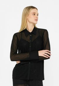 Blouse noire transparente avec un devant à boutons, manches longues, texturée avec des lignes verticales et de petits embellissements, dotée d'un col classique.