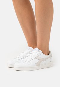 Hvite sporty joggesko med en glatt lærfinish, perforerte detaljer, lyse beige aksenter og flate lisser. Lave design med polstret krage.