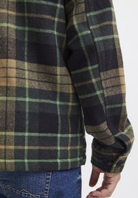 Groen en bruin geruite flanellen shirt met zwarte accenten, met knoopmanchetten en een zachte textuur, weergegeven vanuit de achterkant op een denim achtergrond.