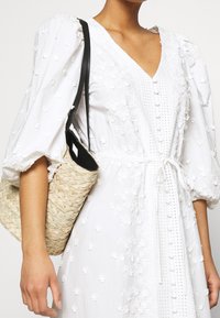 Abito bianco ricamato con maniche a sbuffo, scollo a V e dettagli con bottoni. Abbinato a una borsa di paglia intrecciata con una tracolla nera.