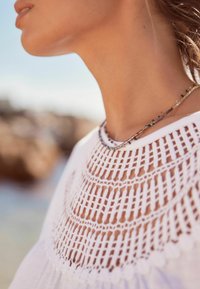 Bijela majica s složenim kukičanim uzorkom oko dekoltea. Ispod se nosi delikatna necklace od više boja.