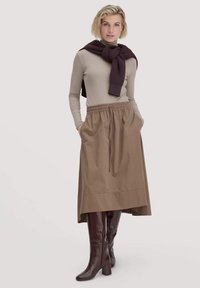 Beige Langarmoberteil kombiniert mit einem braunen, elastischen Rock und burgunderfarbenen knielangen Stiefeln; Schal um die Schultern gebunden.