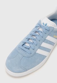 Baskets en daim bleu clair avec trois bandes en cuir blanc, une languette rembourrée et une semelle extérieure en caoutchouc blanc texturé. "GAZELLE" imprimé en jaune.