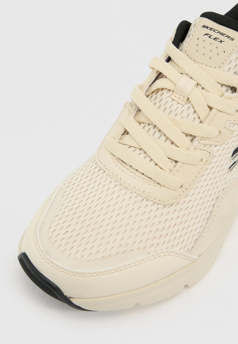 Zapatilla beige claro con parte superior de malla, detalles texturizados y cordones planos. Presenta un collar acolchado y una suela negra con un branding sutil.