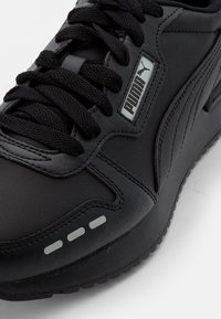 Sneaker sportivo nero con tomaia in pelle strutturata, spesse stringhe nere, etichetta logo argentata e dettagli riflettenti sulla suola intermedia.