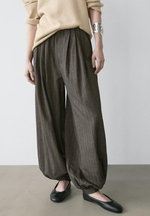 Kobieta w brązowych spodniach w prążki o szerokich nogawkach z mankietami u dołu, beżowym swetrze, srebrnej bransolecie na nadgarstek i czarnych balerinach.