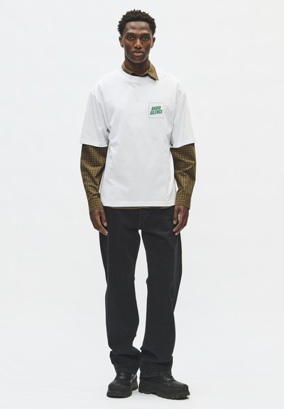 Homme portant une chemise à carreaux marron sous un t-shirt blanc avec le texte "RADIO SILENCE", pantalon noir et bottes noires, debout devant un fond blanc.