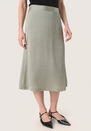 Jupe midi vert clair avec une surface texturée, une taille lisse et une silhouette droite, associée à des escarpins noirs à bout pointu.