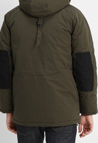 Olivově zelená parka s kapucí, černými loketními záplaty a nastavitelným stahováním. Vyrobena z texturovaného materiálu s volným střihem.
