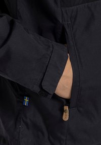Giacca nera con una mano in tasca, etichetta con la bandiera svedese e tirante in pelle sulla zip; tessuto testurizzato e patch in velcro.