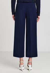 Frau in navyblauen, weit geschnittenen, knöchellangen Hosen, einem navyblauen, kabelgestrickten Pullover und weißen, hochhackigen Slingback-Schuhen, stehend vor einem schlichten Hintergrund.