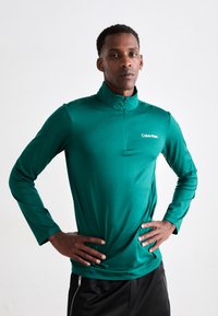 Maglietta verde con zip e maniche lunghe realizzata in tessuto morbido e leggero. Presenta un colletto alto e un piccolo logo sul petto.