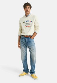 Crèmekleurige trui met kleurrijke nautische graphics, lichtblauwe jeans en gele sneakers. De trui heeft een geribde halslijn en manchetten.