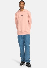 Felpa rosa chiaro con tasca frontale e logo "element" ricamato. Abbinata a jeans blu larghi e scarpe da ginnastica nere.