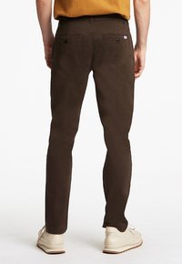 Barna pamut nadrág, slim fit fazonnal, hátul két gombos zsebből áll és sima tapintású. Világos színű cipővel viselve.