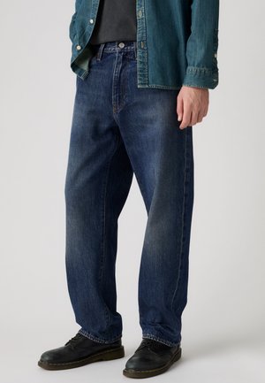 Hombre con vaqueros rectos azul oscuro, zapatos negros, camisa negra y chaqueta vaquera desabrochada, de pie frente a un fondo liso.