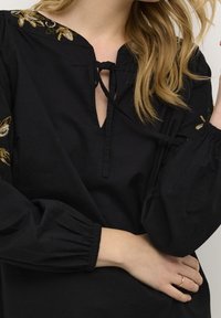 Blouse noire avec un décolleté en keyhole et un détail à nouer. Présente une broderie florale sur les épaules et des manches volumineuses élastiquées.