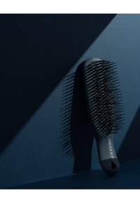 Svart hårbørste med avrundet håndtak, fleksible svarte børstehår og en logo på siden. Bakgrunnen har en mørkeblå gradient.